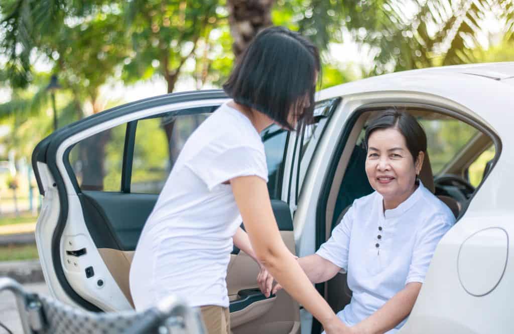 Caregiver Helping Senior Handicapped Asian Women From Wheelchair Get Into Car